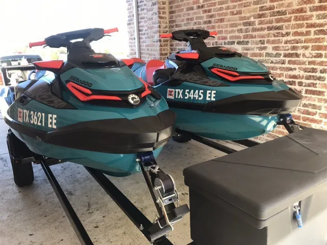 Two teal and black jet skis on a trailer, under a carport with a brick wall.