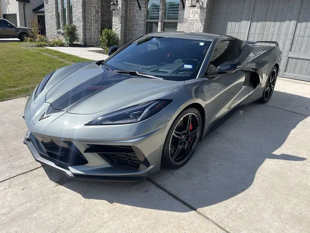 Gray Corvette sports car parked in front of a house with a driveway. Black racing stripe on the hood.